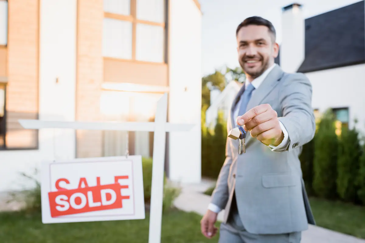 Smiling real estate agent holding house keys and a sold sign after closing a transaction