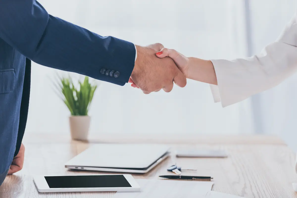 Business broker and client completing a deal with a handshake across a wooden desk representing a successful business transaction