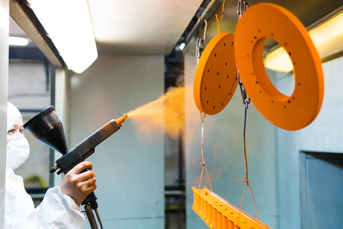 Powder coating technician in a protective suit spraying powder paint onto metal parts in a professional powder coating facility
