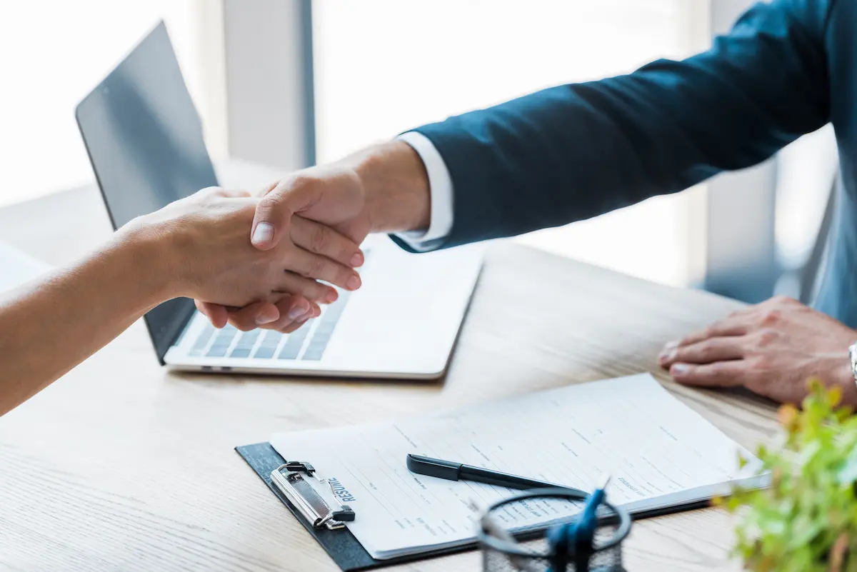 Recruiter and candidate shaking hands with a laptop visible representing the professional staffing and employment placement services that a quality staffing agency provides for employers and job seekers