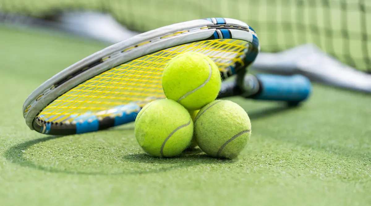 Close-up of a tennis ball and racket on a hard court with a blurred net in the background representing the competitive and recreational tennis experience that a quality tennis association provides for players of all levels