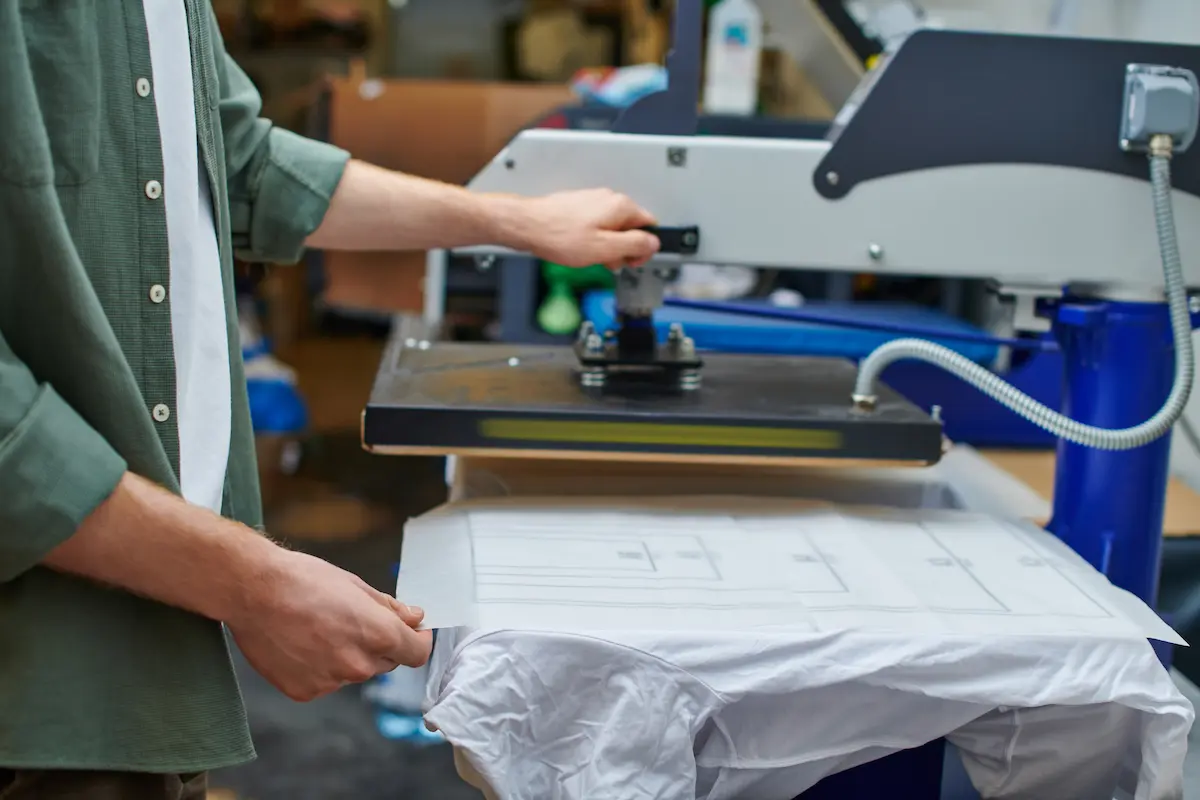 A craftsman holding a printed shirt layer while working at a professional screen printing machine representing the skilled custom apparel production that a quality screen printing company delivers