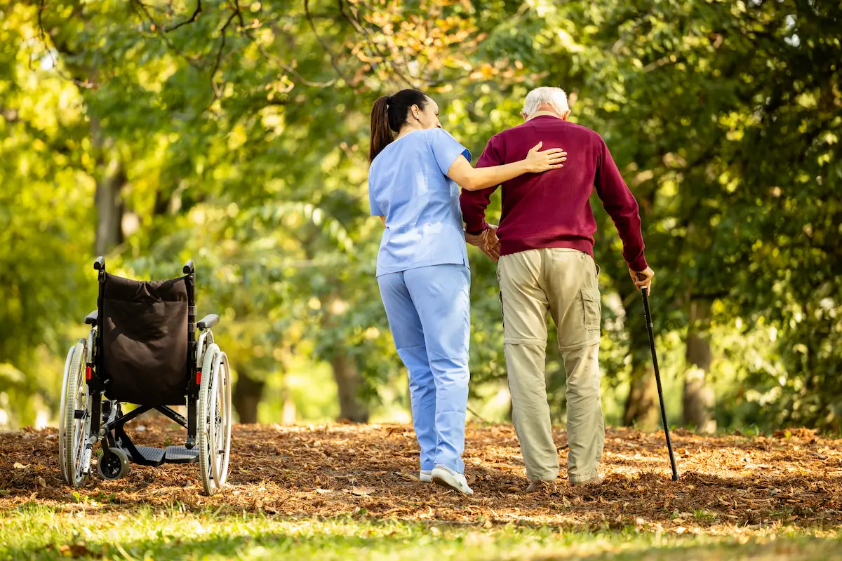 A nursing assistant helping an elderly man walk with a walking cane representing the compassionate and supportive in-home senior care that a quality home care agency provides for seniors and their families