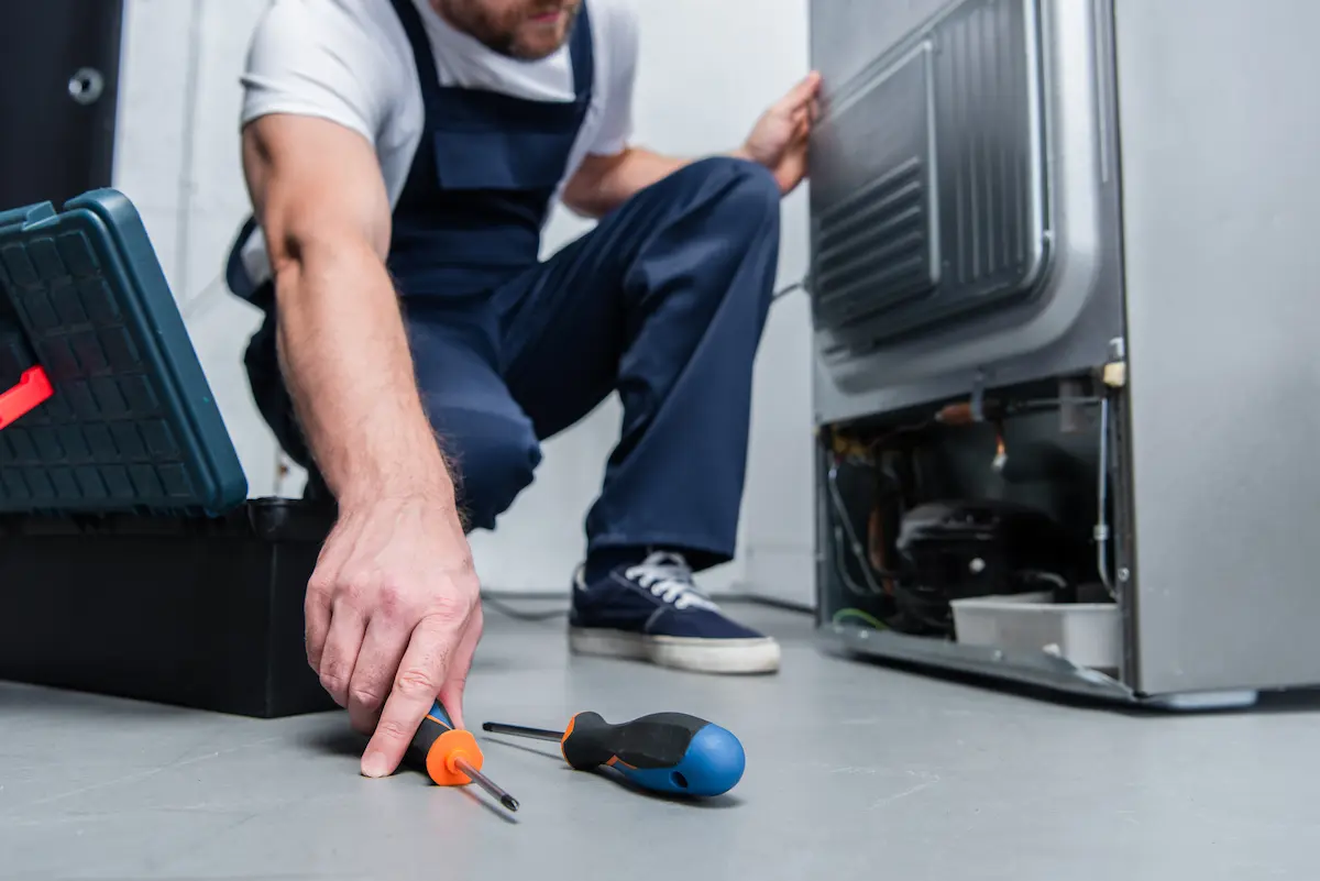A repairman in overalls working on the floor next to a broken refrigerator representing the skilled and professional refrigeration repair service that gets equipment back online fast for homeowners and commercial operators