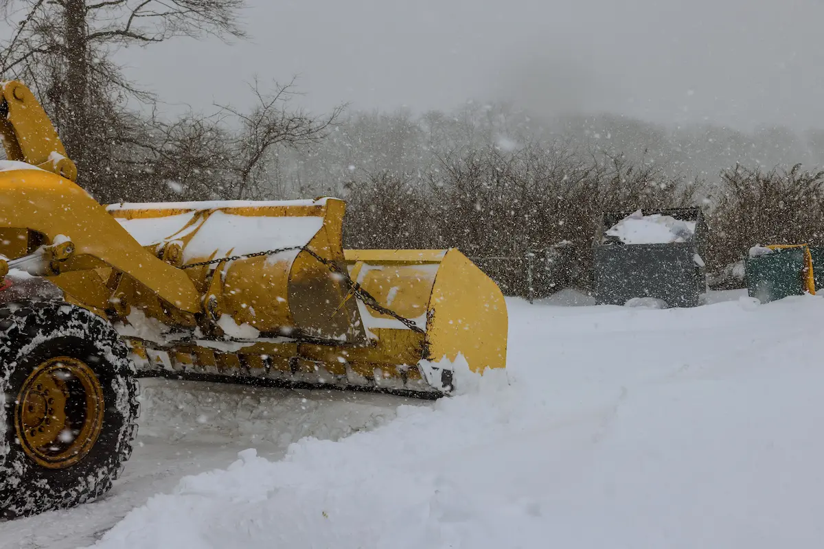 A mechanized tractor used for snow removal parked on a city street during winter snowfall representing the professional equipment and reliable winter property maintenance service that a quality snow removal company provides