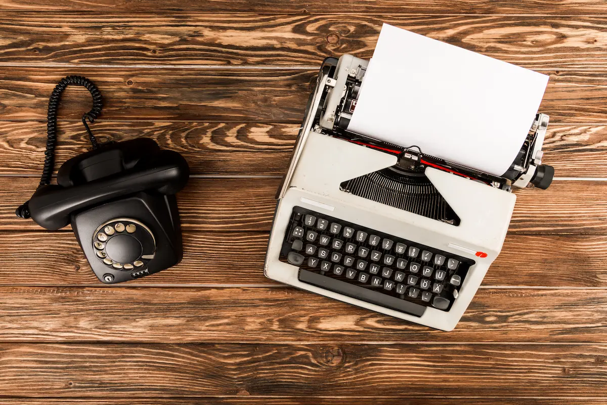A vintage typewriter and rotary dial telephone arranged on a wooden table representing the period props, vintage items, and carefully curated inventory that a professional prop rental company offers to film productions, commercial shoots, event planners, and theater companies