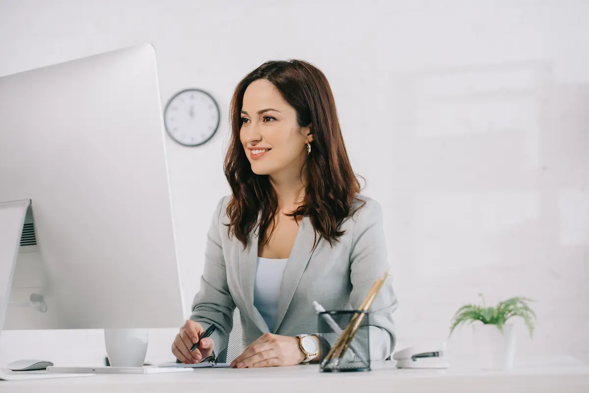 A professional virtual assistant smiling while working at a computer monitor and writing in a notebook representing the organized, efficient, and professional administrative support that a great virtual assistant service provides to small business owners, entrepreneurs, and executives